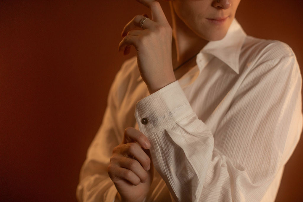 White cotton button up with a tone on tone stripe. Featuring wooden buttons and embroidered latin phrase on the back and cuffs translates to 'finding yourself in the whispers of the world'.
