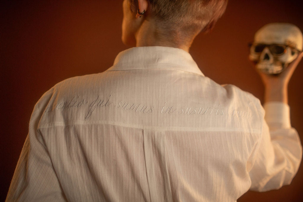 White cotton button up with a tone on tone stripe. Featuring wooden buttons and embroidered latin phrase on the back and cuffs translates to 'finding yourself in the whispers of the world'.