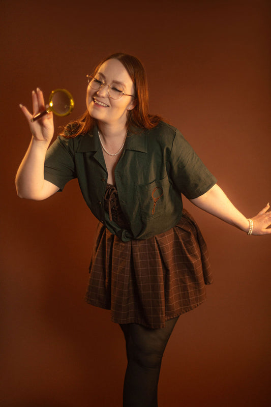 Crisp moss green camp shirt with a linen and cotton blend. With brass brown buttons and a bronze magnifying glass embroidered on the front pocket.