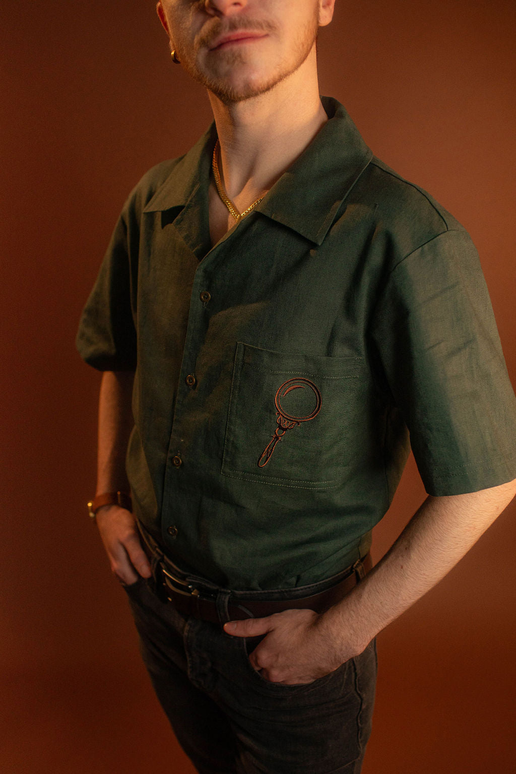 Crisp moss green camp shirt with a linen and cotton blend. With brass brown buttons and a bronze magnifying glass embroidered on the front pocket.