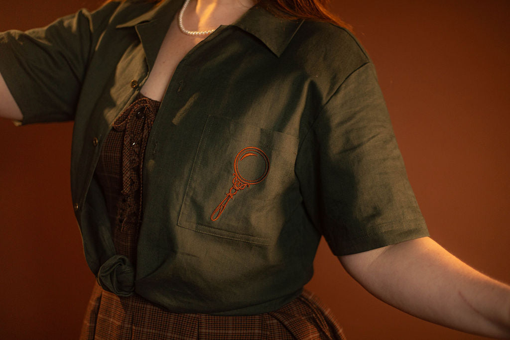 Crisp moss green camp shirt with a linen and cotton blend. With brass brown buttons and a bronze magnifying glass embroidered on the front pocket.