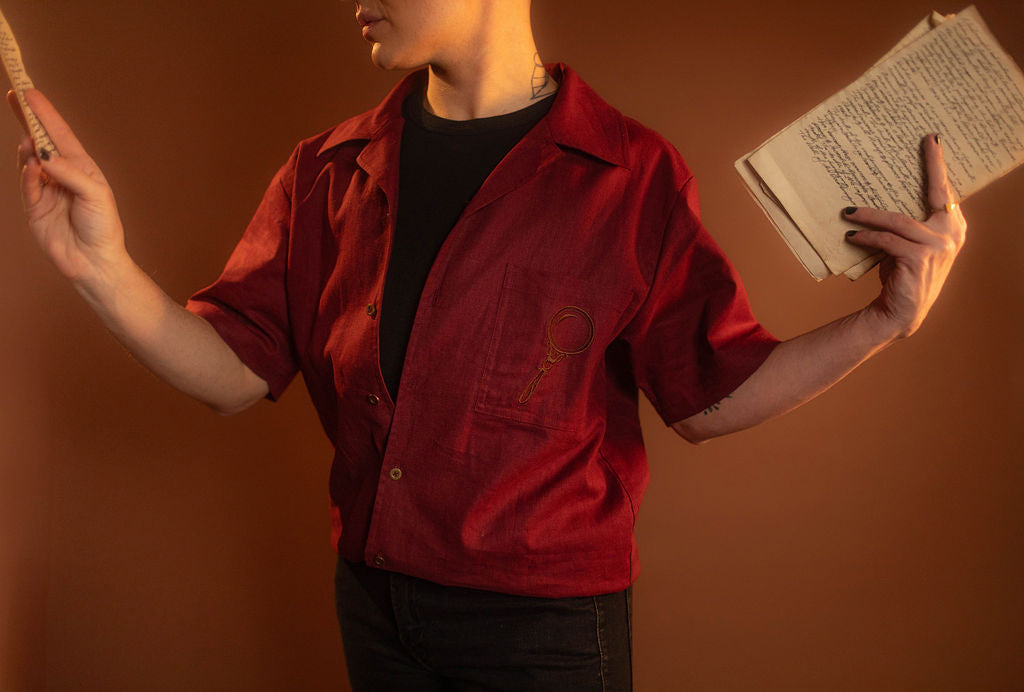 Crisp wine red camp shirt with a linen and cotton blend. With brass brown buttons and a bronze magnifying glass embroidered on the front pocket.