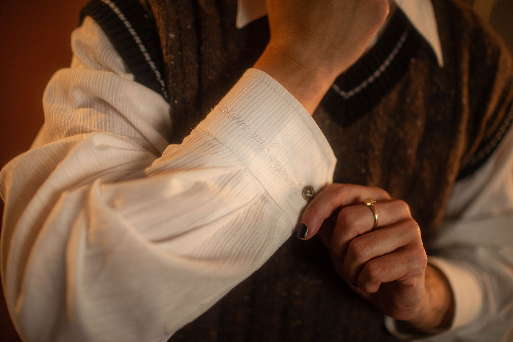 Brown and black wool and nylon blend tweed sweater vest with tennis sweater style black and off-white striped collar and sleeve cuffs. A small, black embroidered magnifying glass on front chest. Featured with our Classics White Button up.