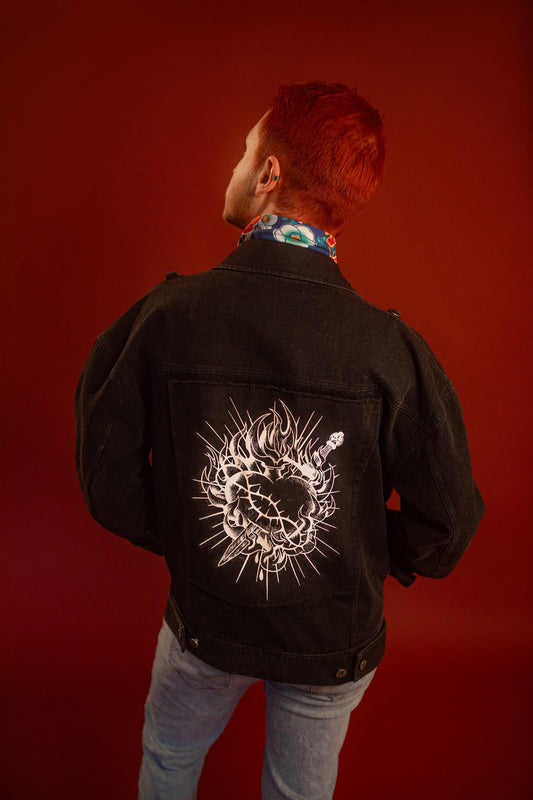 Black sturdy cotton denim jacket with white screen-printed sword on chest pocket and sacred heart graphic on a back patch. Custom pearl button on front pocket aligned with sword's pommel, five pockets, and multiple reinforced D rings for clipping accessories on to, like our Heart On Your Pocket Chain. Shown here with our Lovers Favor Bandana.