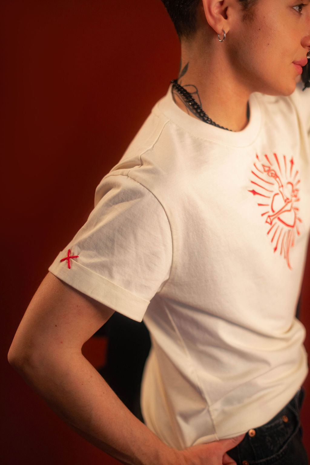 Off-white 100% cotton tshirt with embroidered red sword and heart design on chest, cuffed sleeves with a hand-tacked red X on one, printed collar information, and Saints & Sonnets heart and sword logo on a red hem tag and printed in red on the back collar.