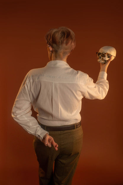 White cotton button up with a tone on tone stripe. Featuring wooden buttons and embroidered latin phrase on the back and cuffs translates to 'finding yourself in the whispers of the world'.