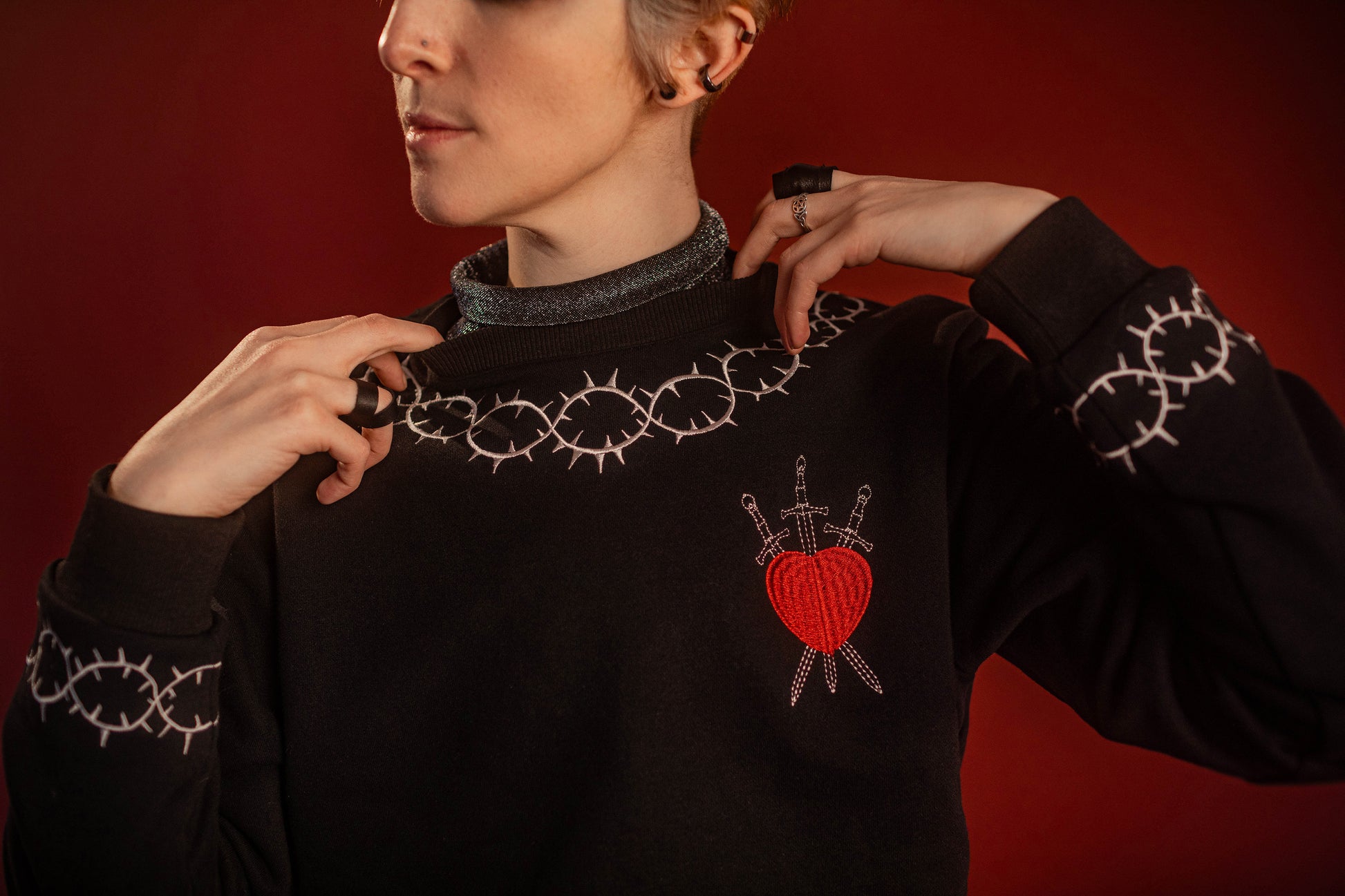 Soft 100% cotton fleece-lined black crewneck sweatshirt. Soft-backed embroidered silver thorn pattern around the collar and cuffs, silver and red three of swords embroidered on chest, with three swords crossed between a red heart. Featured here with our Chainmaille removable Collar dickie.
